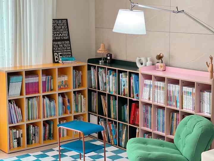 Living room reading space featuring yellow, deep green, and pink bookshelves with a green armchair and warm ambient lighting
