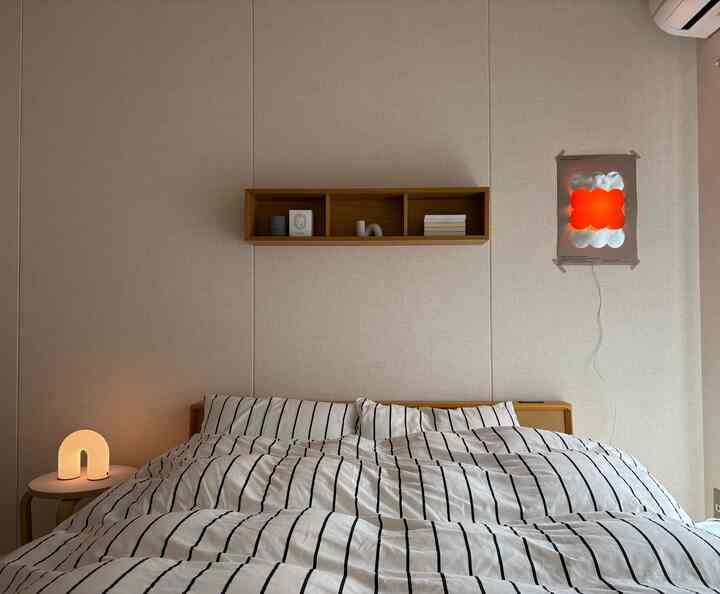 ホワイトと木目調のシンプルな寝室、ストライプの寝具と壁棚、温かみのある照明が配置された雰囲気あるインテリア