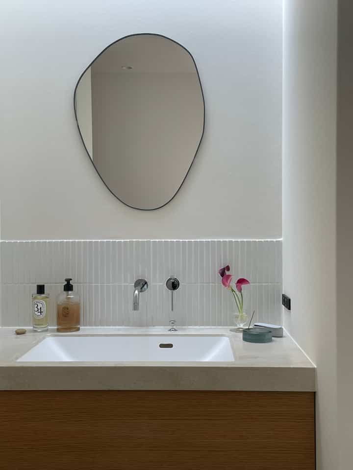 白とブラウンのトーンのバスルーム洗面台、不規則な形の鏡とピンクの花が活けられた花瓶が配置されたシンプルな空間