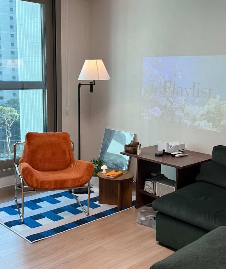Natural tone living room with orange armchair, dark green sofa, floor lamp, and projector setup creating a modern and cozy atmosphere