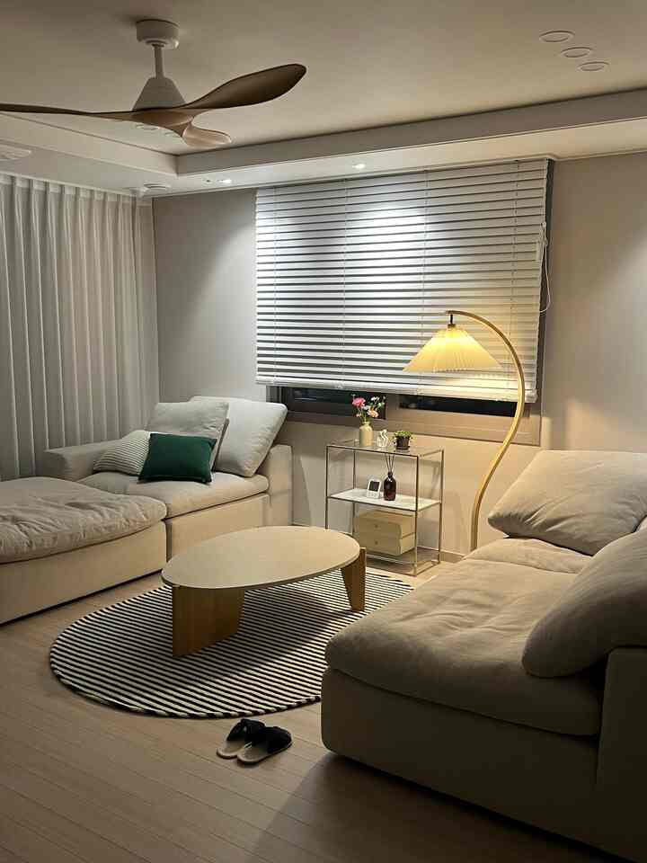 白とベージュの色調のリビング、L字型のベージュのモジュラーソファと楕円形のコーヒーテーブル、曲線的なフロアランプが配置された印象的な空間