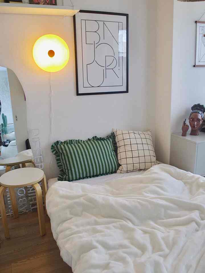 ナチュラルカラーとウッドトーンの一人暮らし寝室、ホワイトのベッドとグリーンのクッション、壁掛けライトとアートが調和した温かみのある空間