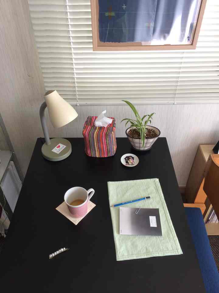 黒いテーブルと白いブラインドがあるコンパクトなワンルームのデスクスペース、テーブルランプと植物、カップが配置された雰囲気のあるインテリア