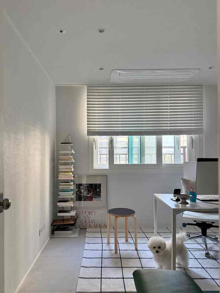 A compact home office space in white and gray tones featuring a bookshelf, small wooden stool, and a small white dog with clean decor