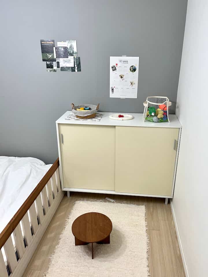 グレーの壁とナチュラルカラーの子供部屋、左下に子供用ベッド、中央に小さな丸いテーブル、右にペットのおもちゃが置かれたキャビネットが配置された空間