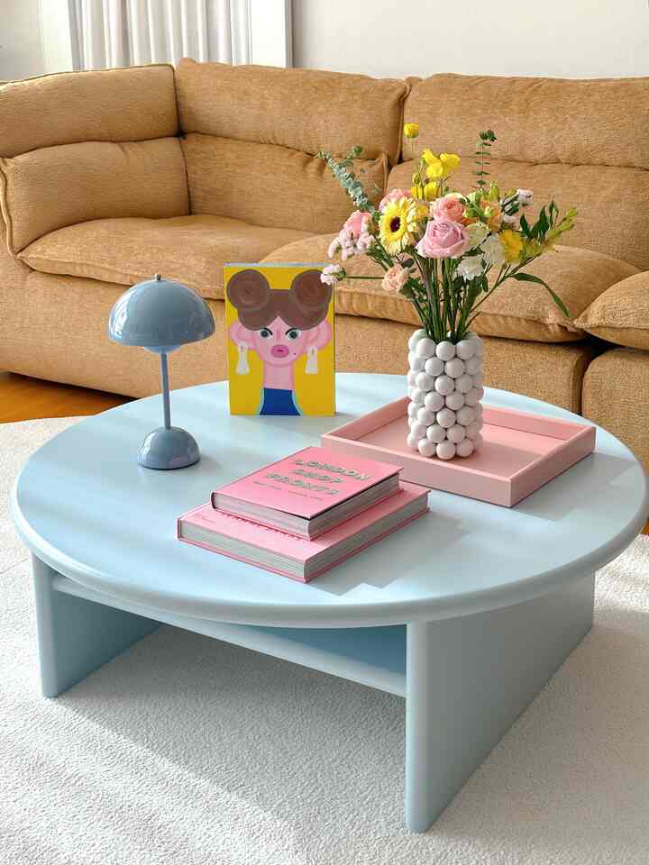 Pastel-toned living room featuring a brown sofa, sky blue round sofa table, pink accessories, and a vase with fresh flowers in a bright cheerful ambiance