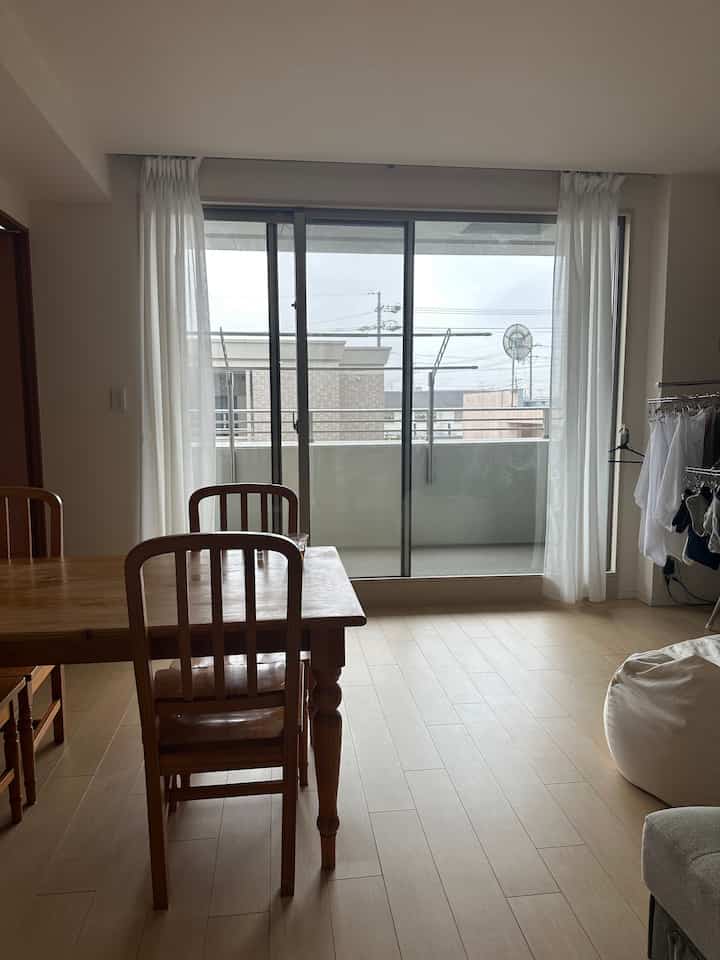 明るい木目調の床とウッドダイニングテーブルがあるナチュラルモダンなリビング、白いカーテンと大きな窓ガラスが配置された空間