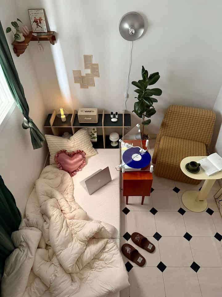 White and brown toned small hobby room featuring bed, checkered sofa, turntable, and side table in cozy arrangement