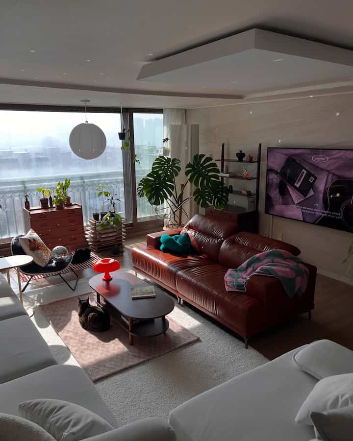 A monotone living room featuring a warm camel leather sofa and white sofas, complemented by natural light and indoor plants creating a cozy atmosphere
