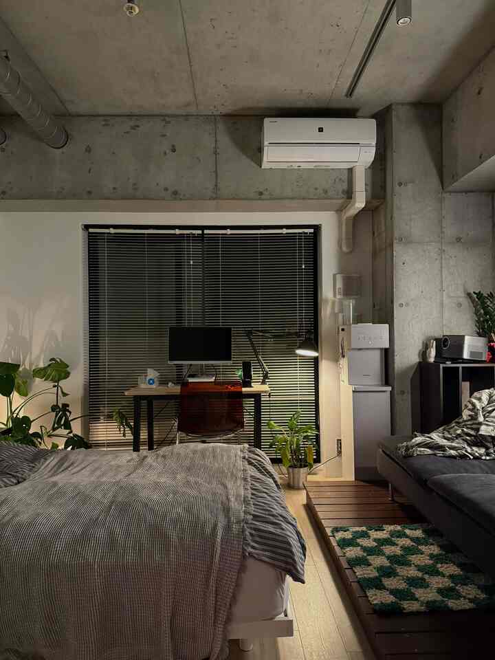 Natural wood-tone floor and concrete walls in a studio apartment featuring bed, desk, and sofa creating cozy single household space