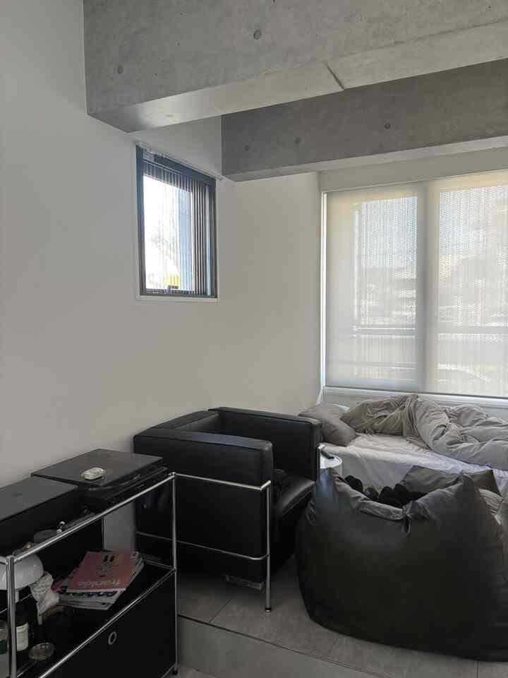 白い壁と露出コンクリートの天井が印象的なモダンなワンルーム寝室、黒のアームチェアとビーズクッションが配置された落ち着いた空間