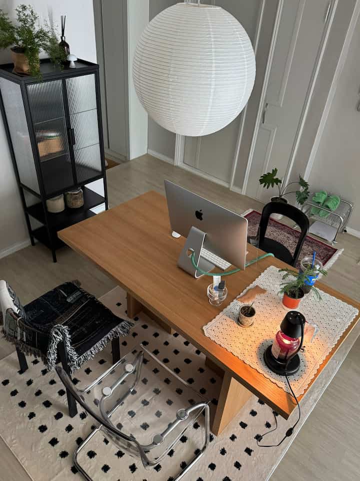 Natural wood-toned home office space featuring a solid wood table, transparent chair, pendant lighting, and plants in a tidy cozy interior