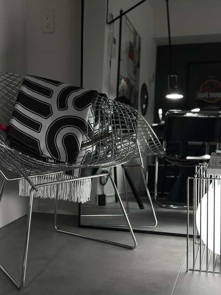 グレーの床と黒い壁を背景に、シルバーのワイヤーフレームチェアと金属製ケージランプが配置されたモダンでインダストリアルなリビングルーム