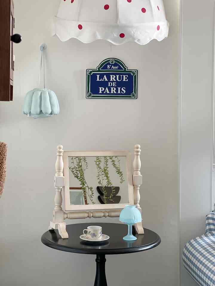 白い壁と黒い丸型サイドテーブルがあるフレンチカントリースタイルの空間、クリーム色の木製フレームの鏡とパステルカラーのテーブルランプが配置されている