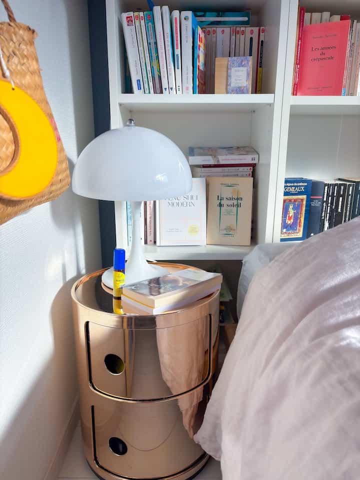 白とゴールドの色調の寝室コーナー、ゴールドのチェスト、白いテーブルランプ、本棚が配置された雰囲気のある空間