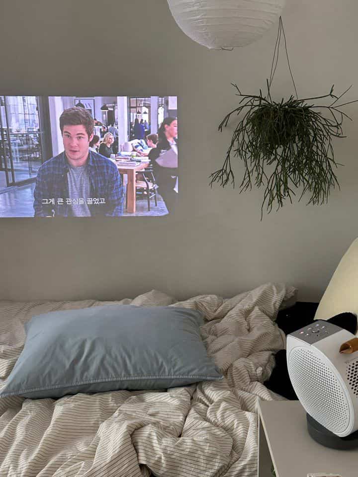 モノトーンのベージュとグレーの寝室、広いベッドと枕、壁のプロジェクタースクリーンと吊り下げられた植物が配置された落ち着いたインテリア