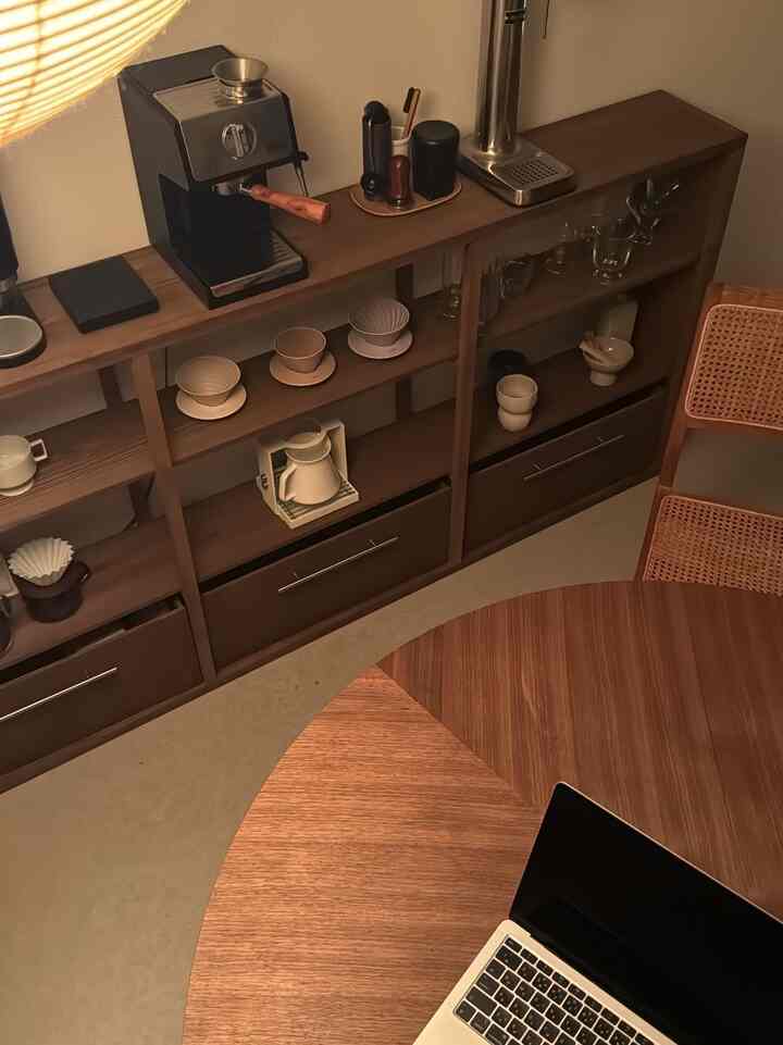 Natural wood-toned Nordic living room featuring shelves with handle storage boxes, a coffee machine, rattan chair, and a round table in warm ambiance