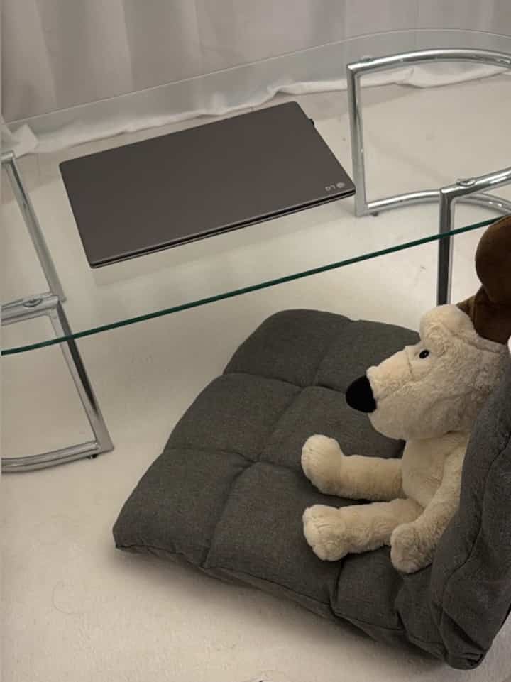 ホワイトトーンのワークスペース、グレーのクッションとぬいぐるみが配置され、ガラステーブルにノートパソコンが置かれている