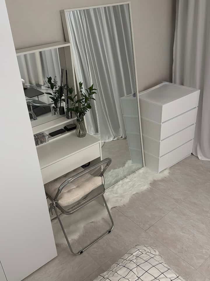 白を基調とした空間、大きな全身鏡とチェスト、透明な折りたたみ椅子と化粧台が配置されたシンプルでモダンなインテリア