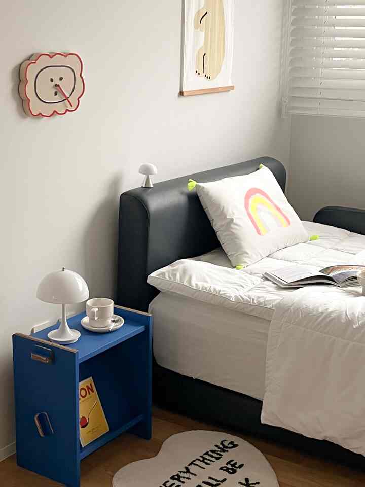 A white and blue toned bedroom featuring a black bed, blue side table, small mushroom-shaped lamps, and a colorful cushion in a simple modern setting