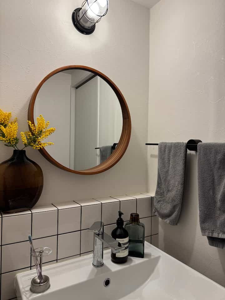 白い壁とタイル、丸い木製フレームの鏡が配置された清潔感のある洗面台のある浴室
