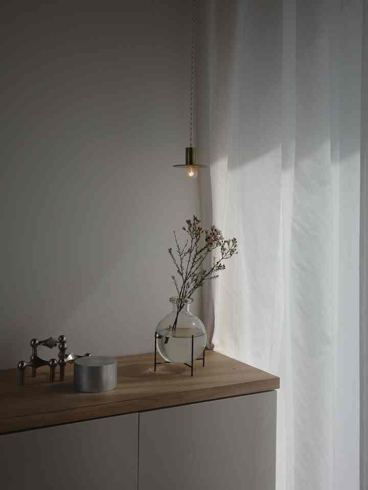 柔らかな自然光が差し込む白いカーテンと木目調のキャビネット上に花瓶とペンダントライトが配置された玄関スペース