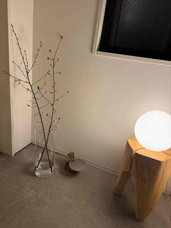 ナチュラルカラーの玄関、透き通った花瓶に桜の枝が挿され、木製スツールの上に丸い照明が置かれたミニマルなインテリア