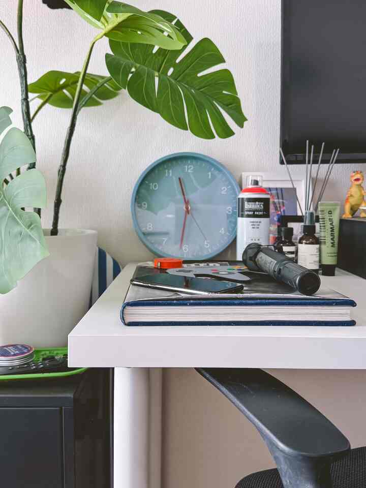 ナチュラルな色調のワンルーム、白いデスクの上にブルーの時計とモンステラのフェイクグリーンが配置された雰囲気のあるホームオフィス空間