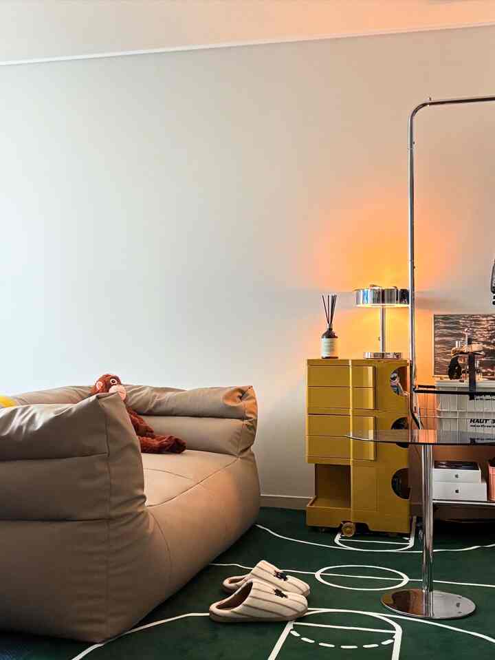 A cozy small living room with a beige bean bag and green patterned rug, featuring a yellow storage cart and warm table lamp lighting