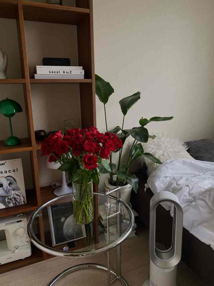 白い壁とベッドがあるモダンな寝室、中央のガラステーブルに赤いバラの花束と植物が配置された雰囲気のある空間