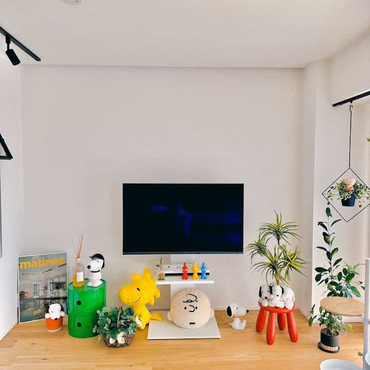 白い壁と木の床のリビングに、テレビ台、多彩なキャラクターのぬいぐるみ、植物、赤いスツールが配置された印象的な空間