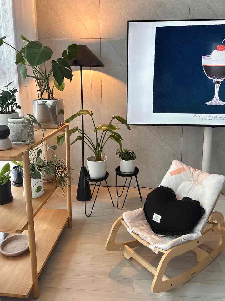 A cozy living room with wood tones and black accents, featuring wood shelves, various plants, and a cute cushion on a chair