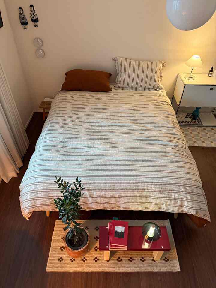 Natural beige-toned bedroom featuring a bed frame with striped bedding centrally placed, a side table with lamp beside it, creating a simple atmosphere