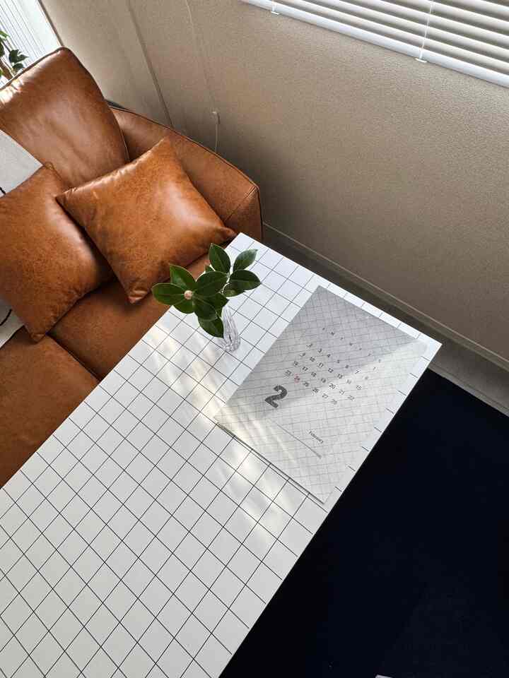 ブラウンのソファと青いタイルカーペットが配置された、窓のブラインドから自然光が差し込む居心地の良いリビングルーム