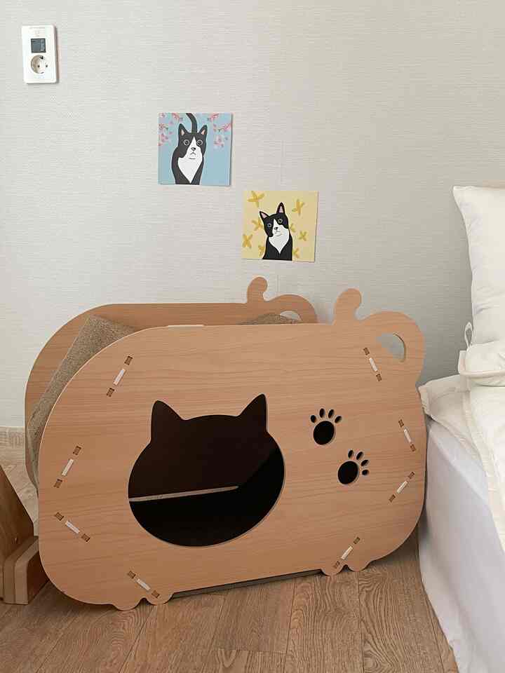 ベージュ色の寝室の壁に猫のイラストが飾られ、木目調の猫型の爪とぎ家具が配置された雰囲気のある空間