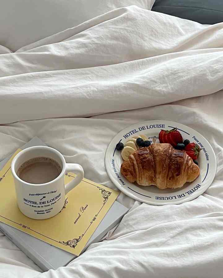 白い寝具の上にクロワッサンとフルーツの皿、コーヒーのマグカップが置かれた雰囲気のある寝室のおうちカフェ