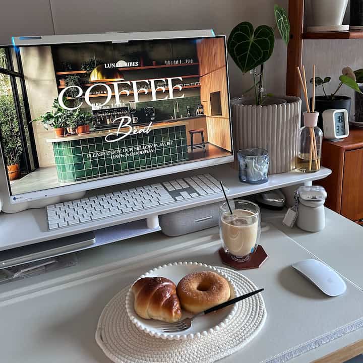 明るいホワイトトーンのホームオフィスのデスクにコーヒーとパンが置かれ、植物やディフューザーが配された雰囲気のあるおうちカフェ空間