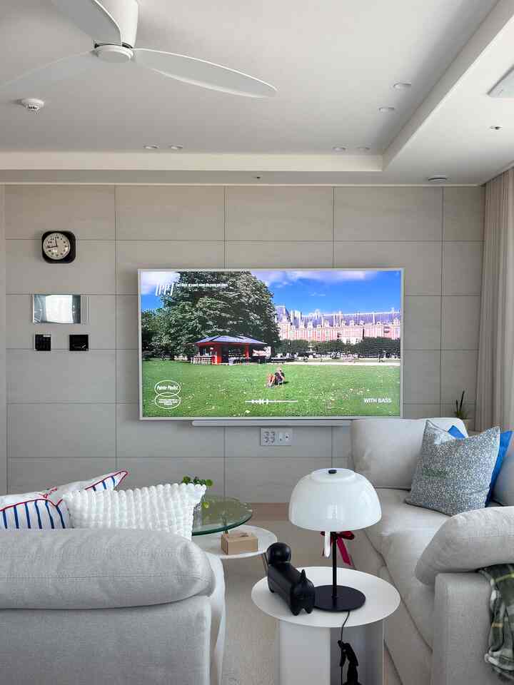 White and beige toned living room featuring large TV, modern sofas, and table lamp creating a cozy atmosphere