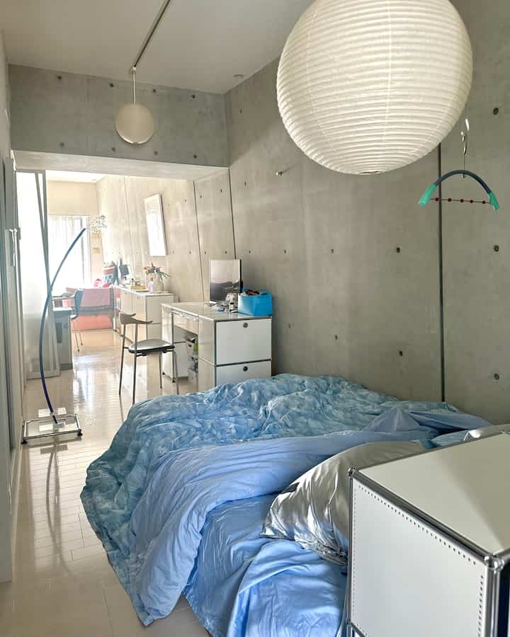 ブルーの寝具とコンクリート壁のある狭くミニマルな寝室、ホワイトのペンダントライトとドレッサーが配置された空間