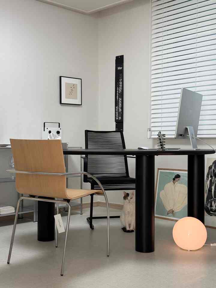 白い壁とブラインドのあるモダンなホームオフィス、黒いデスクと椅子、床には猫と照明が置かれた雰囲気のあるインテリア