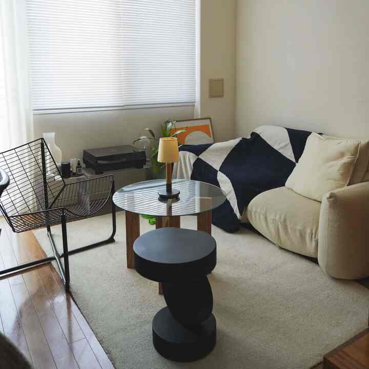 ベージュのソファと黒のスツール、ガラステーブルがあるナチュラルな雰囲気のリビング、窓のブラインドで自然光を調整