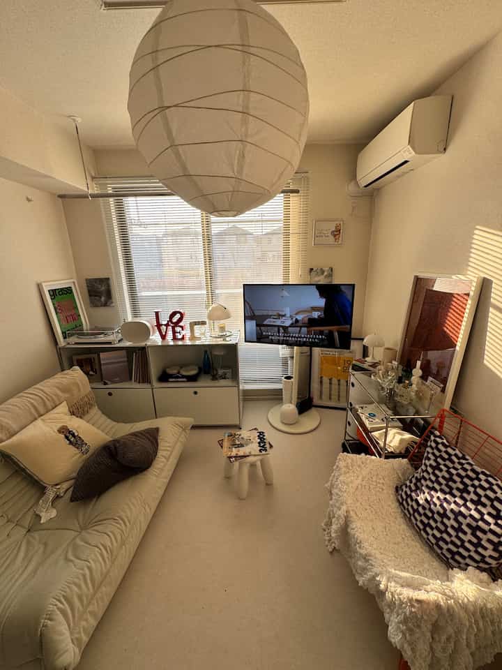 Cozy compact living room in warm beige and white tones, featuring a central pendant light, sofa, and TV stand