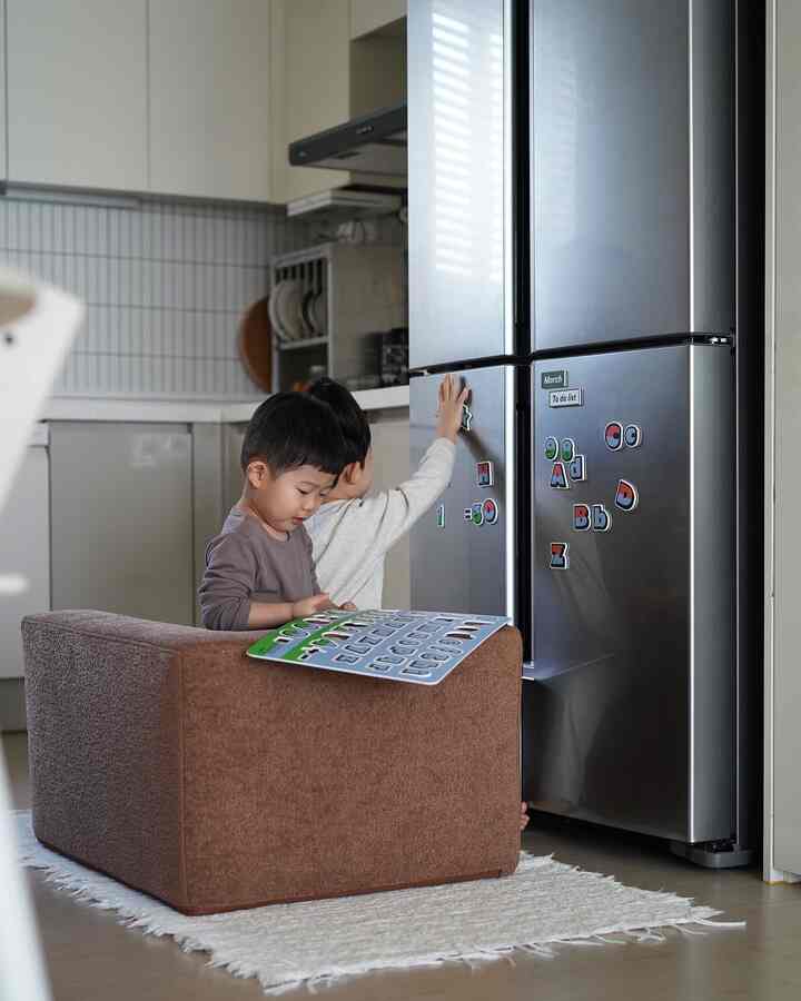 柔らかな自然光が差し込むキッチン、子供用ソファと冷蔵庫のそばで遊ぶ子どもたちがいる温かみのある家族空間