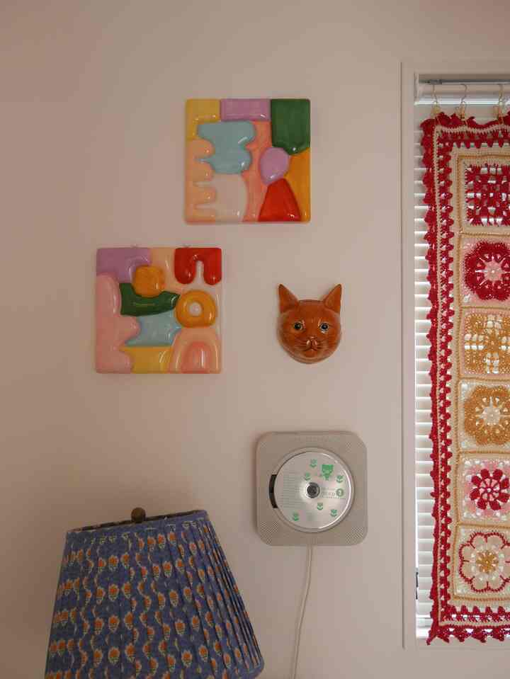 柔らかい色調のリビングの壁に、多彩な陶器のウォールアートと猫の顔の装飾、青いランプシェードと編み物のカーテンが調和した雰囲気のあるインテリア