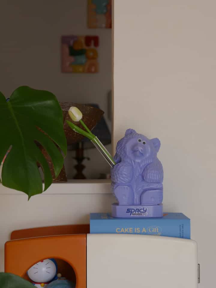 A colorful storage book box and plant arrangement in front of beige wall featuring a lavender bear objet on a blue book