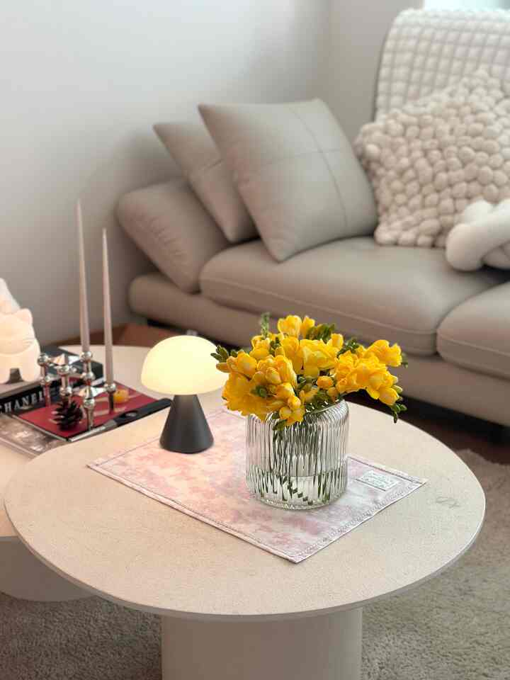 淡いグレーのソファとベージュの円形テーブルがあるリビング、黄色のフリージアの花とミニ照明が配置された雰囲気のあるインテリア