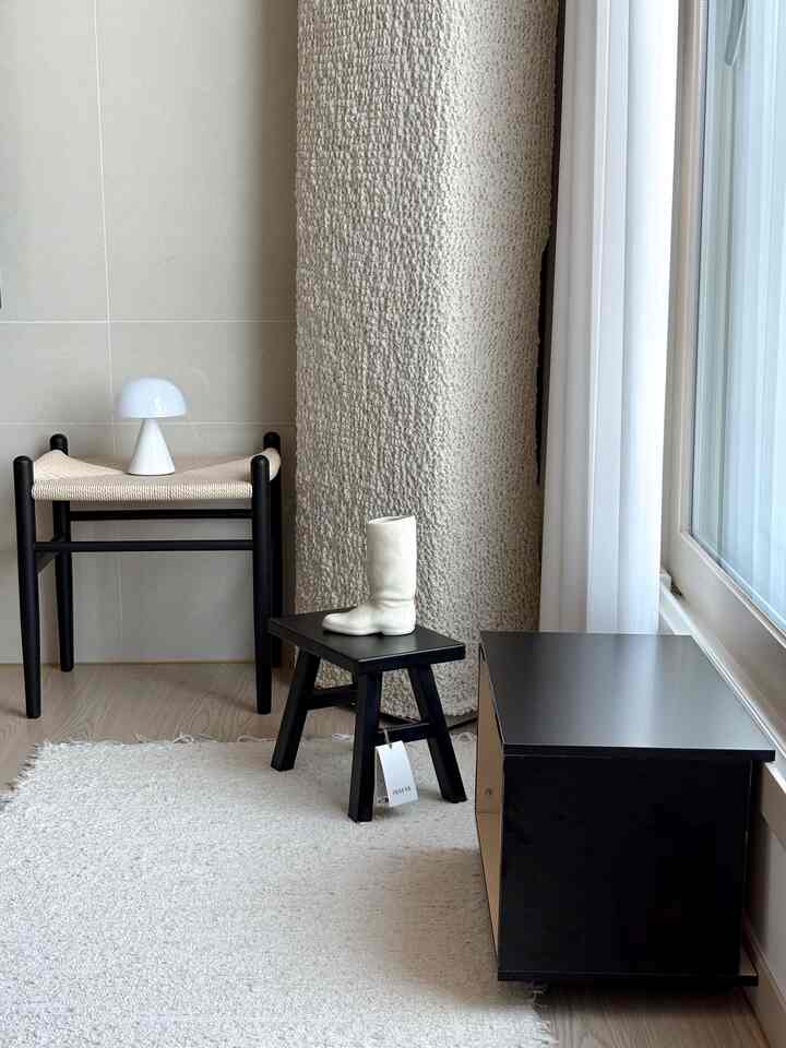 白いカーテンとベージュのラグのある小さな玄関スペース、黒いスツールと収納が配置されたシンプルでモダンな雰囲気のインテリア