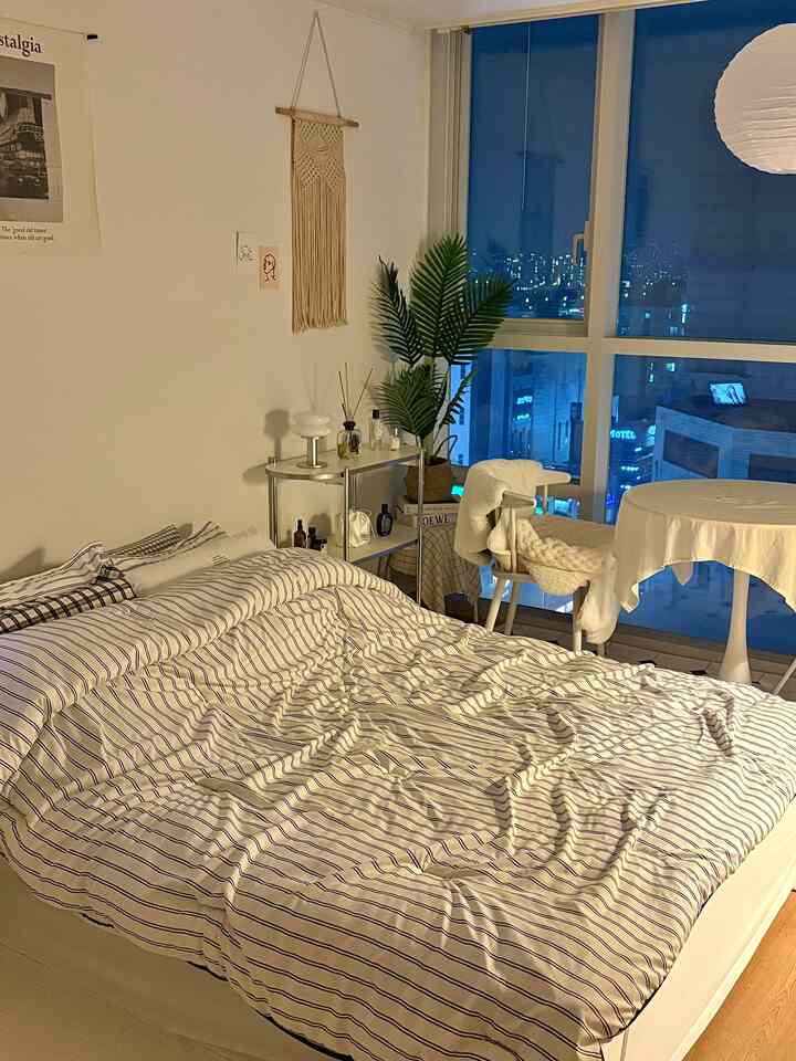 ホワイトとナチュラルトーンのワンルーム寝室、ストライプの寝具とマクラメのウォールハンギング、大きな窓から漢江の夜景が見える温かみのあるインテリア