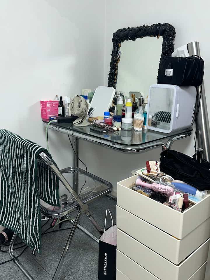 透明な椅子と化粧品が置かれた黒いフレームの鏡の前に白いトロリーが配された狭い部屋のシンプルな書斎兼化粧台スペース