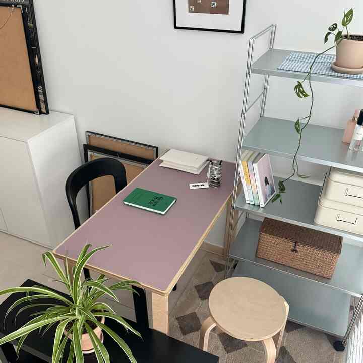 白い壁とグレーの棚があるモダンな部屋、紫色のテーブルマットが敷かれた机と木製スツール、植物が配置された印象的なホームオフィス